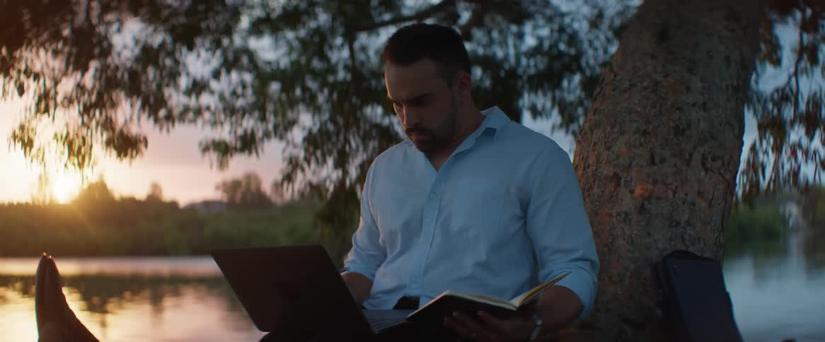Man sitting under a tree writes on a notebook while using a laptop
