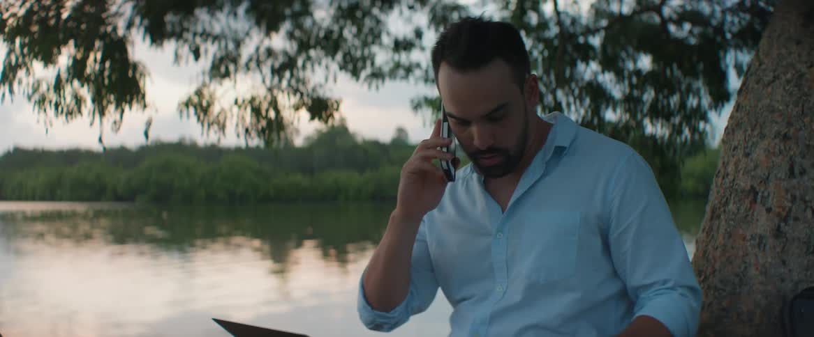 A man sit under a tree working on a laptop and talking on phone