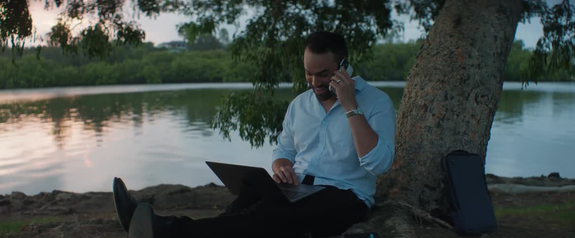 An adult man sits next to a tree facing a river while talking on his cell phone and using his laptop