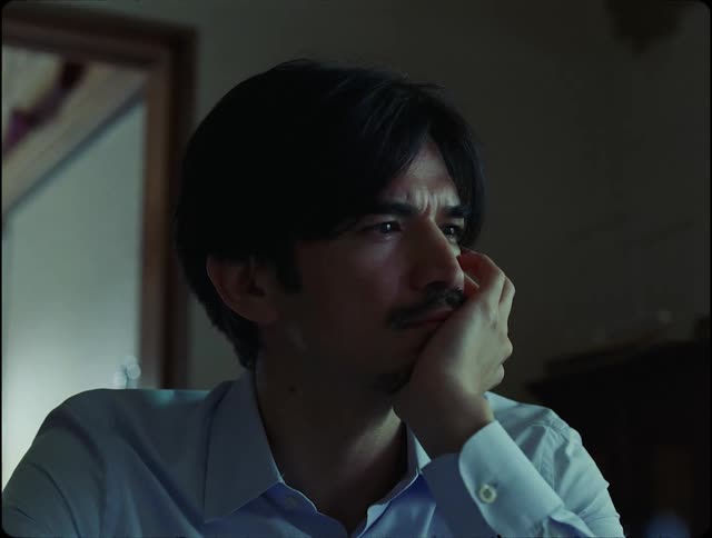 A man with a mustache and a blue shirt sits deep in thought with his hand on his chin