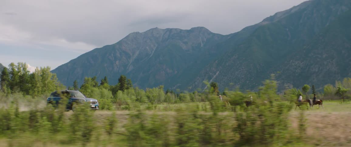 People on road trip pass people riding horses across field bordering mountains