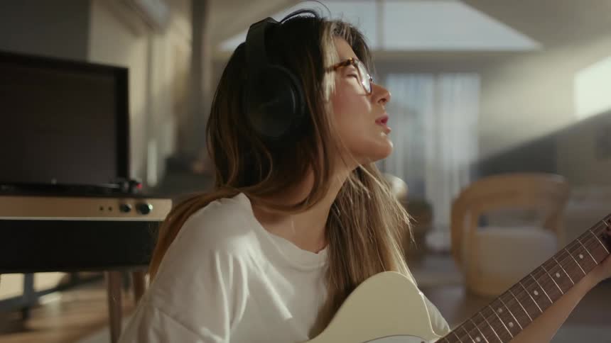 A woman with brown hair wearing a white tshirt and headphones plays a guitar in the living room while singing
