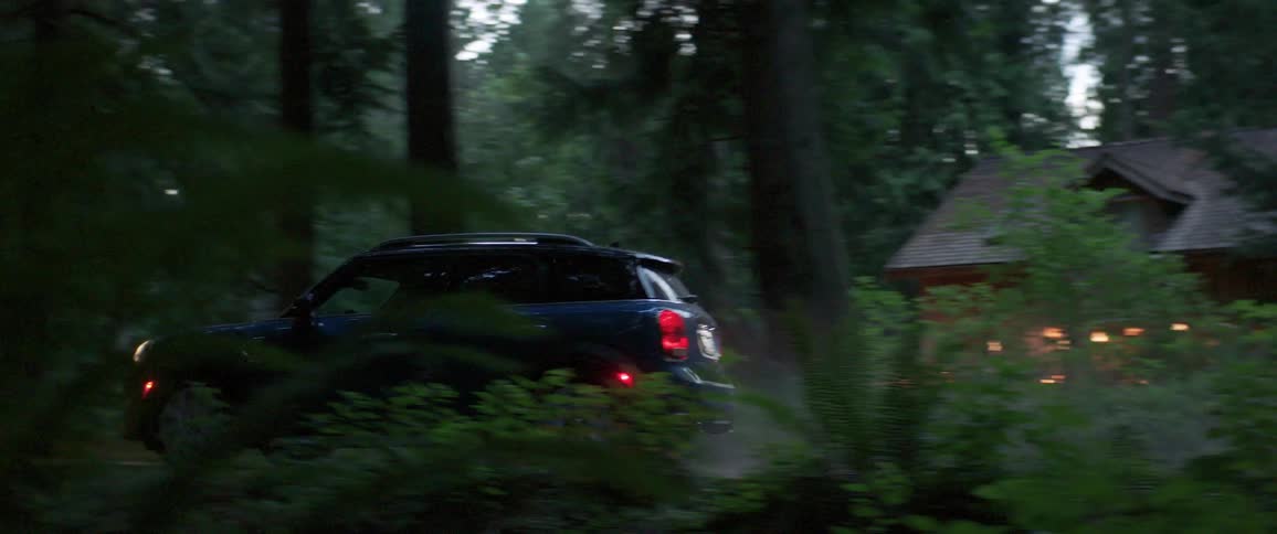 A blue car drives on a country road passing by a cottage in the woods