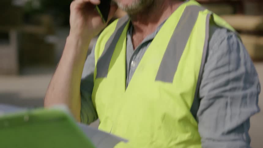Worker standing outdoors at lumberyard consults clipboard during phone conversation