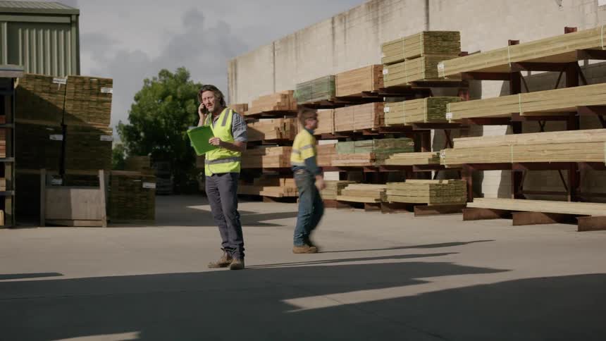 Worker standing outdoors in lumberyard speaks on phone