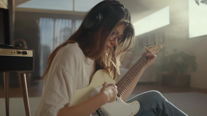 Lady plays guitar indoor with headset on while sitting on a center rug