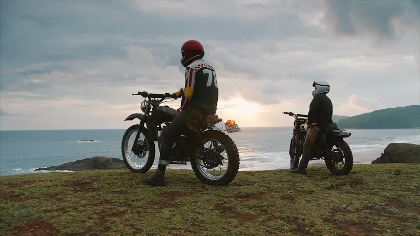 Two motorcyclists sitting  atop a grassy hill overlooking the ocean on a cloudy day
