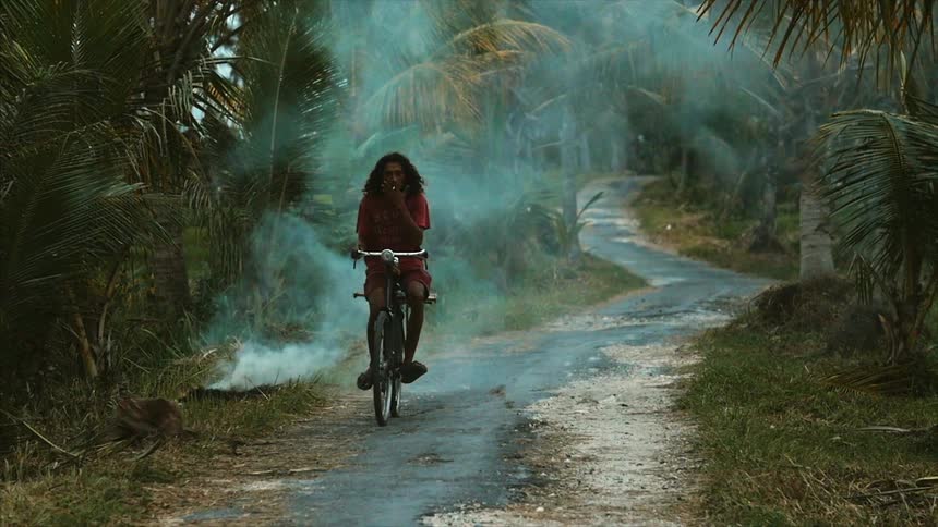 A man riding a bicycle along a road while a small fire burns on the roadside
