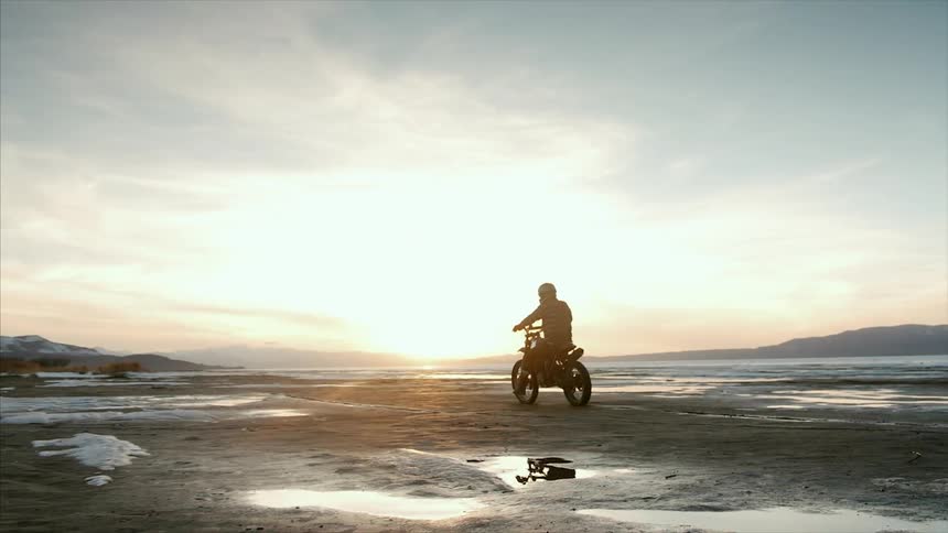 A man rides his motorcycle across a large field at sunset
