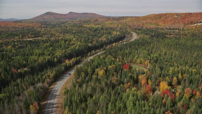 Traffic moves on a road through a forested residential rural area with trees in bright fall colors and forested mountains in the distance