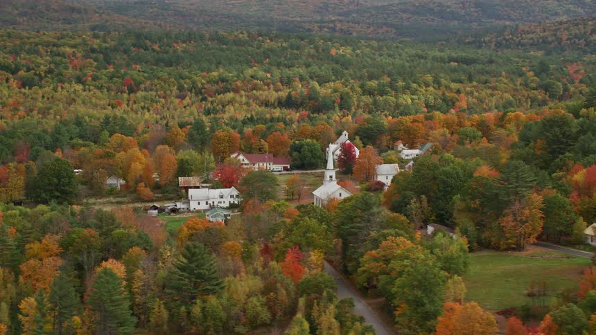 A small village with a white church in the middle of a large forest with some trees changing the color of their leaves to orange
