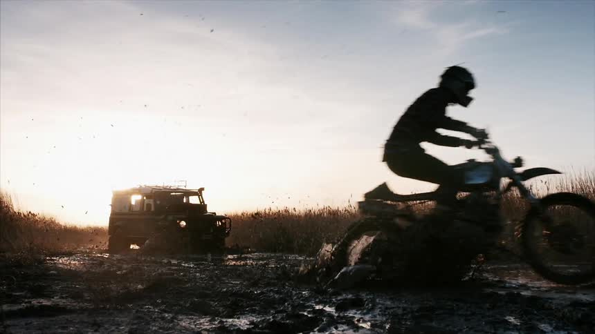 A truck drives through the mud at sunset as a dirt bike passes by