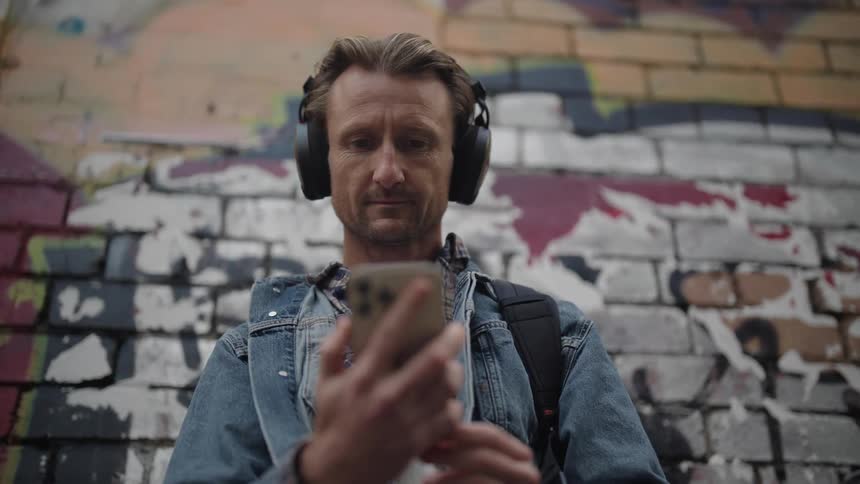 A man putting on overear headphones in front of a graffiti wall while checking his cell phone