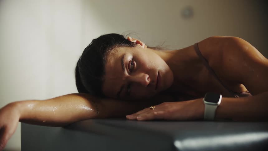 Sweaty woman with sports bra and a silver wristwatch rests on black chair