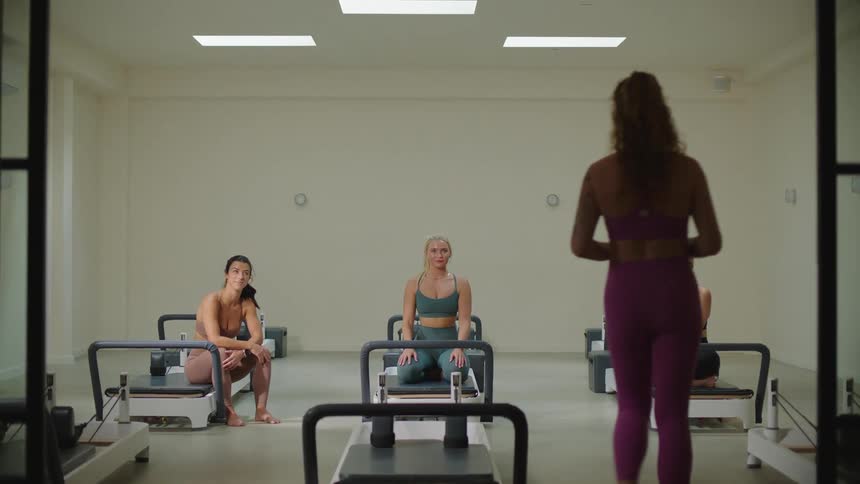 Three woman are seated on pilates reformer machines in a spacious exercise studio while woman walks in front giving instructions to women