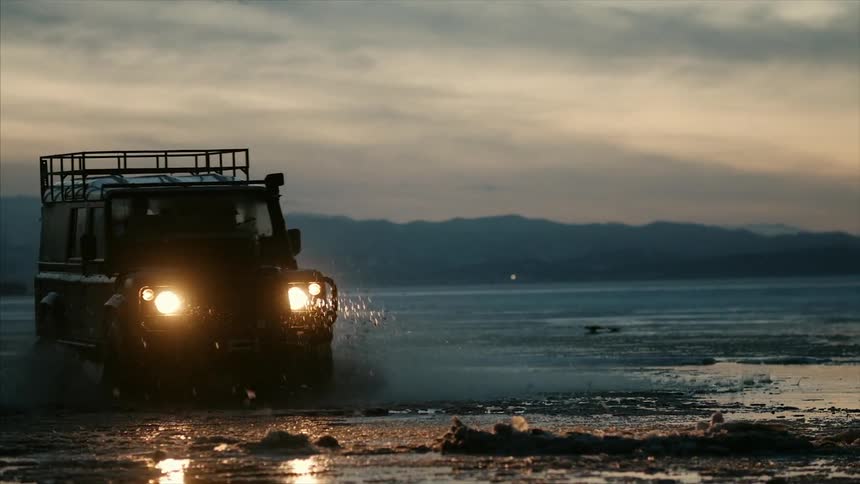 A large suv driving through water with mud flying in the air at sunset