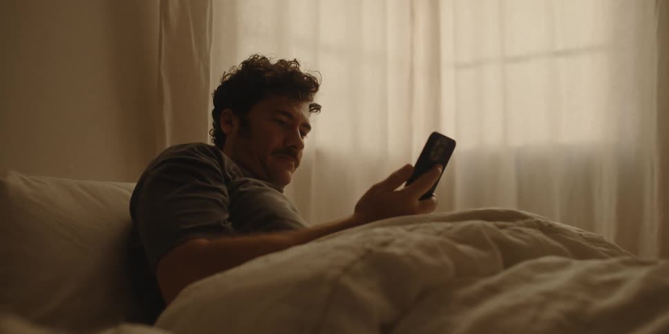 A man leans over while in bed to retrieve his smart phone