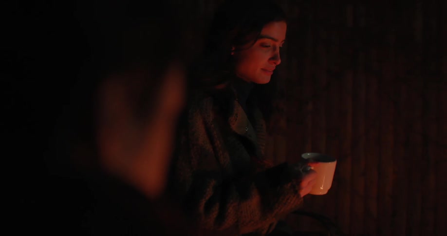A woman by a campfire at night drinking from a mug and smiling