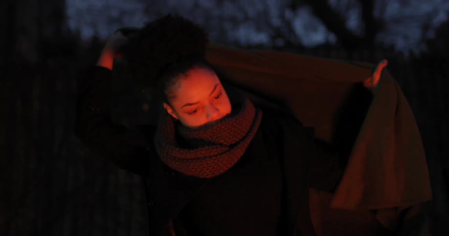 Woman places blanket over shoulders as she stands beside a fire