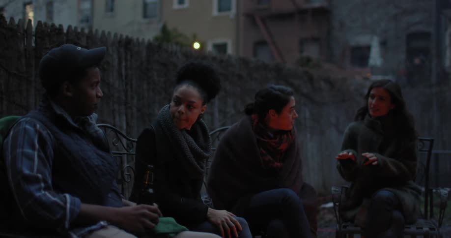 People sit in a yard talking by a campfire