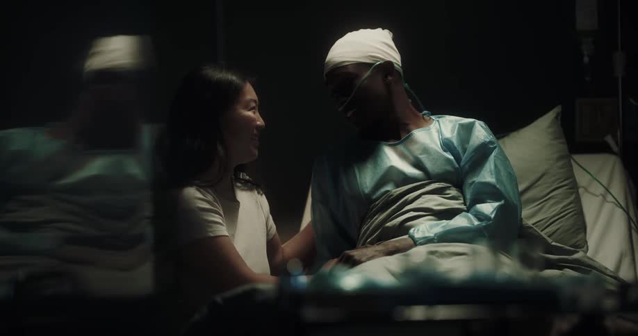 A woman sits by her boyfriend in a hospital bed as they talk and laugh