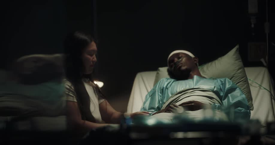 A woman reads a book and affectionately tends to a man with a nasal cannula lying in a hospital bed