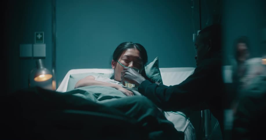 Asian lady drinks from a glass offered by the black man seated beside her hospital bed