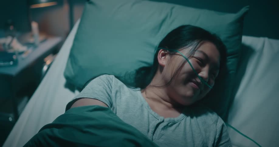 Asian lady drinks from a glass offered by a black man seated near her hospital bed