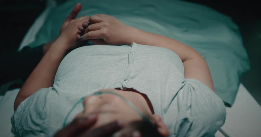 Sick lady lays on the hospital bed while a black man rubs her head and hands