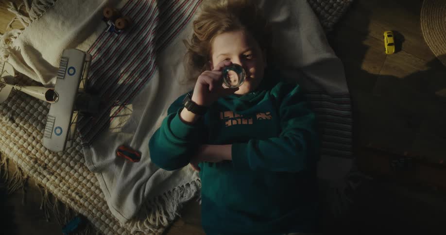 A little girl lying on the bed is observing a glass lens with her eye