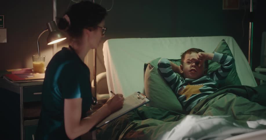 Nurse checking a child patient lying in bed taking their temperature and making notes