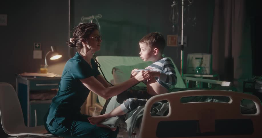 A nurse uses her stethoscope to check the heart of a boy on a hospital bed as he tries playing around
