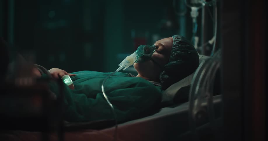 A woman wearing a hospital gown lies in a hospital bed her eyes closed with an oxygen mask on her face