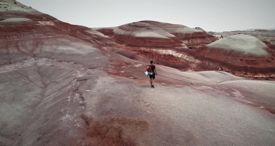 From above a man in sunglasses a red tshirt and black shorts hikes a hill overseeing a desert landscape