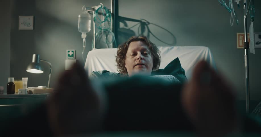 A woman is lying in a hospital bed and tries to move her feet but cannot and makes expressions of anguish