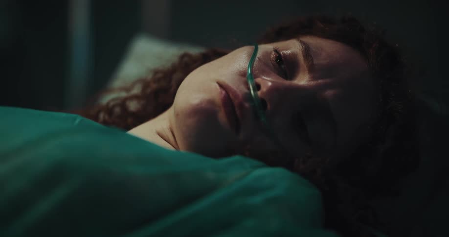 A woman lies still in a hospital bed with a sad look in her eyes