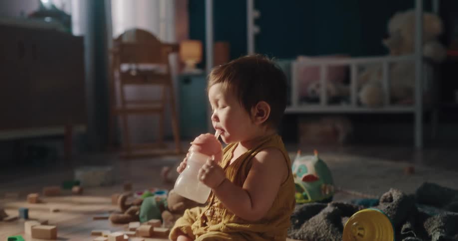 Baby girl drinking water and then starting to crawl on a floor filled with toys in a room