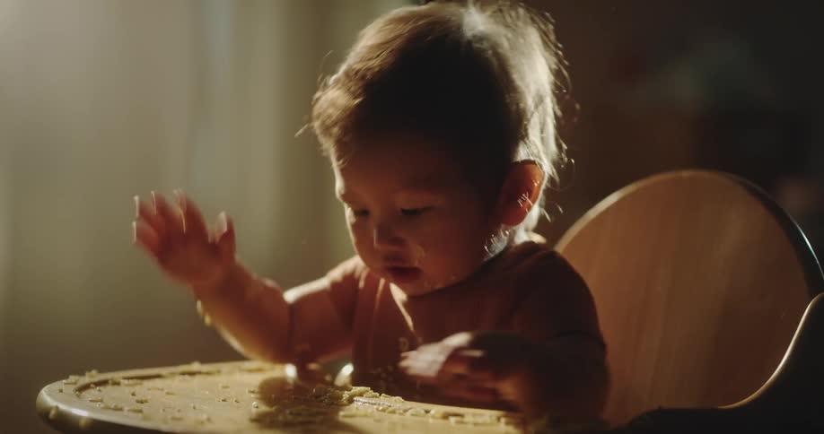 A baby in a highchair plays with their food breaking into pieces