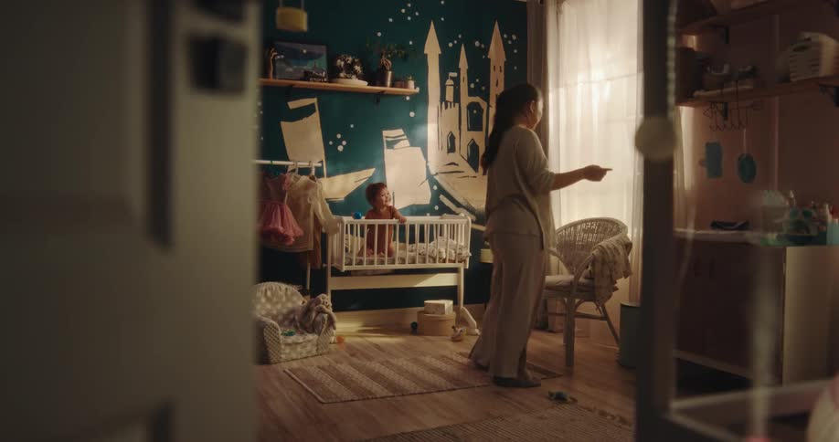The mother is kneeling beside of the bed that the child is sitting in and then she gets up and walks to do something