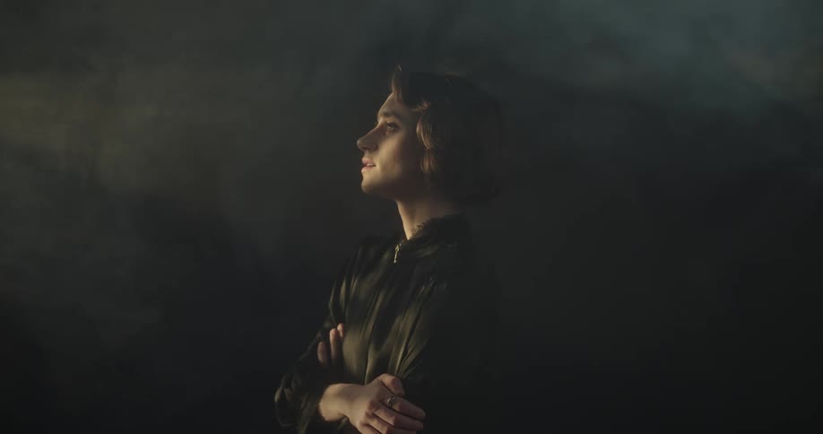 A woman stands sideways against a black background with passing light reflection