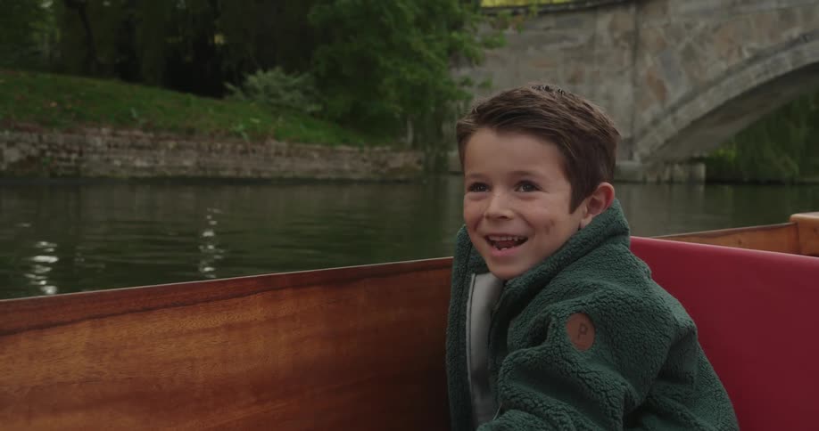 Boy riding in boat nearing bridge smiles while looking at water