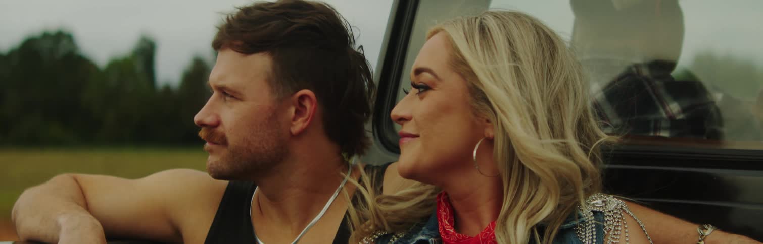 Man and woman sit in back of truck driven through countryside by man