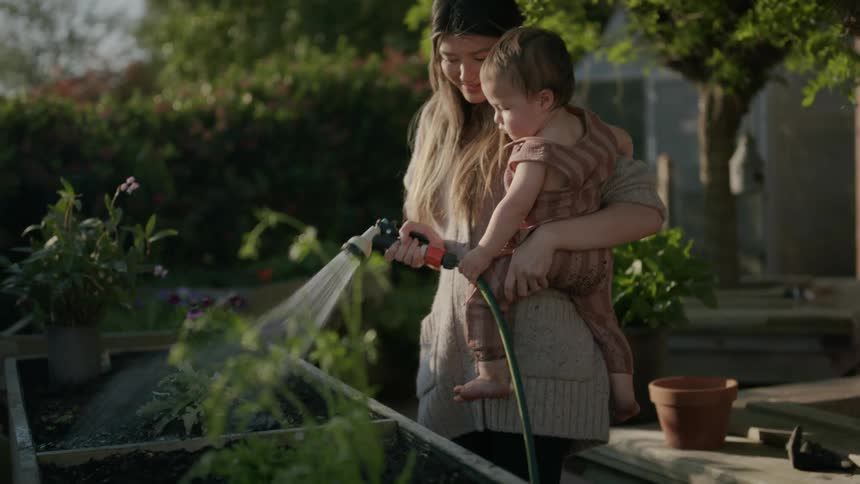 Woman holds toddler while water garden with hose