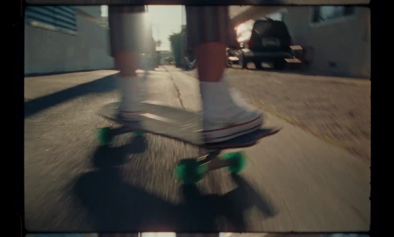 A person is riding a skateboard on a street with some cars parked in the gutter