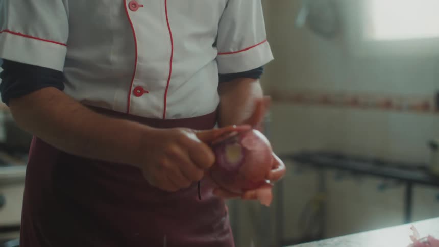 A person cutting an onion with a knife and on the table there are several carrots