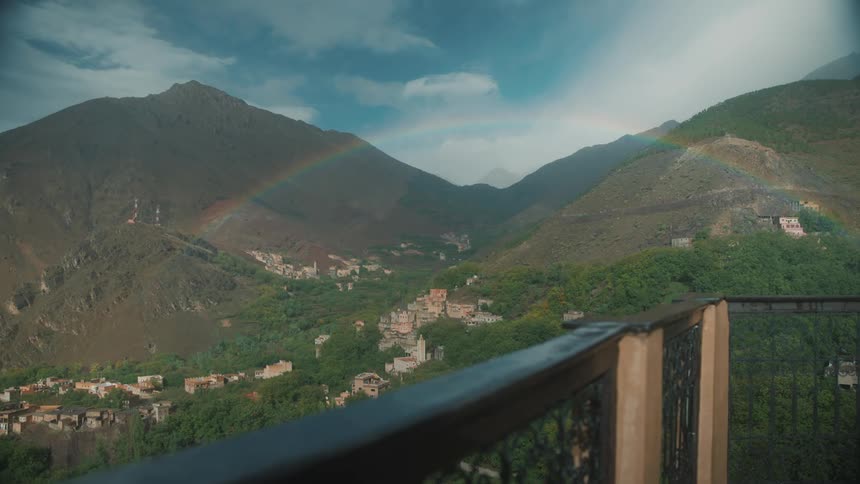 A balcony overlooking a mountain village with a rainbow in the sky