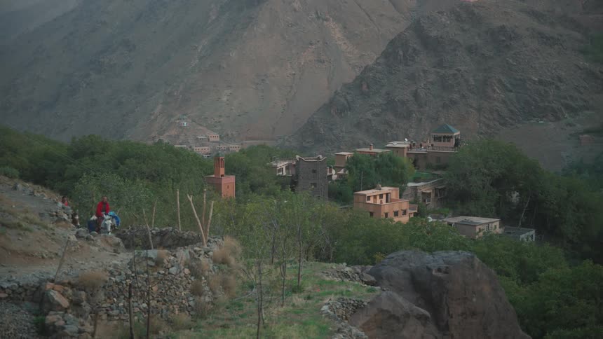 Two people riding horses up a hill on a trail with a village nestled among the mountains on the horizon