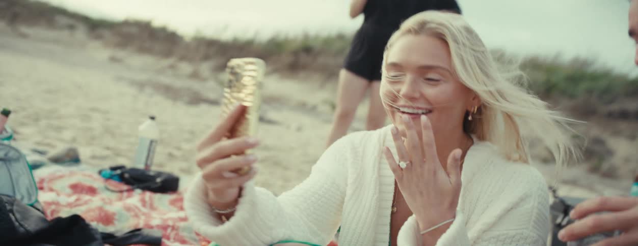 Newly engaged couple sitting on beach share news via video call