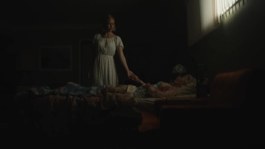 A young woman wearing a dress is standing in a dark bedroom by a bed holding her dying grandmothers hand