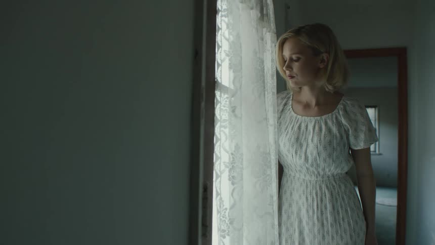 A woman with blonde hair wearing a white dress stands next to window curtains